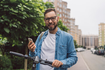 Portrait of a young handsome smiling casually dressed man posing outdoors with mobile phone in hands while walking with his electro scooter © wpadington