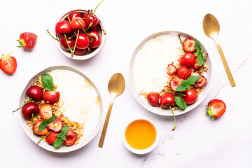 Greek yogurt with granola and fresh strawberries, cherry and honey, decorated with aromatic mint leaves on white marble background top view. Healthy tasty homemade breakfast concept. 