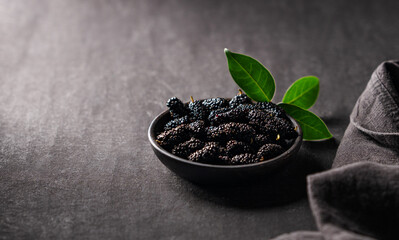 Fresh black mulberry with leaf in black wooden bowl on dark background with napkin. Concept of juicy and healthy seasonal garden berries. Copy space.