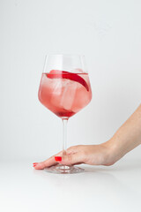 A refreshing pink cocktail in a wine glass, garnished with a slice of fruit and ice cubes, is held by a hand with red nail polish. The minimalist white background emphasizes the vibrant drink.