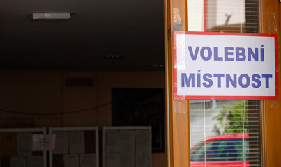 A paper sign with the Czech inscription meaning "Polling room" stuck on the open entrance door to the building on election day in the Czech Republic, boards with voting ballots in the background