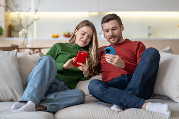 Cheerful happy couple sitting on sofa with smartphones, smiling, playing absorbing online game, watching short funny videos in internet, exchanging photos, having group video call with friends