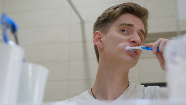 A young guy brushes his teeth with a toothbrush in front of the mirror in the bathroom