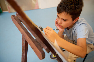 Young boy focused on drawing in art class, creating artwork on an easel