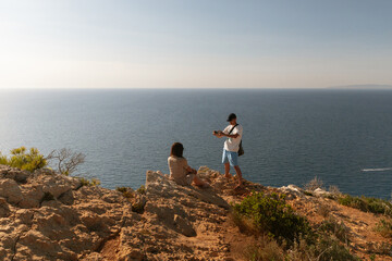 A young guy takes a photo of a girl on his phone by the sea.