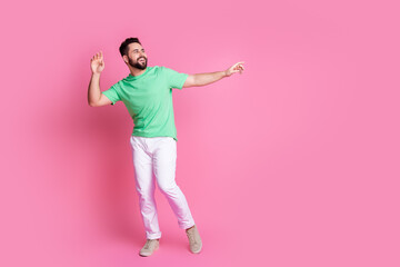 Full size photo of carefree handsome guy dressed green t-shirt white pants look empty space dancing isolated on pink color background