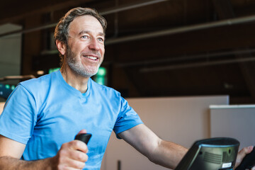 Fototapeta premium A senior man is smiling while riding a exercise bike in a gym.