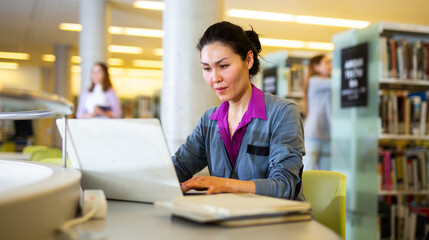 Interested Asian woman sitting at table in library, using laptop, books and taking notes for research. Self-education concept..