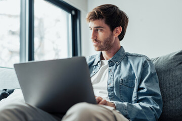 Dreamy pensive freelance man thinking over work project ideas on home office couch, using laptop, looking at window away, smiling at good thoughts, future business vision..