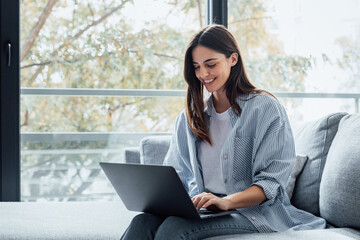 Concentrated millennial girl sit on couch working on laptop browsing internet at home during lazy weekend, focused young woman freelancer busy using computer surfing wireless web shopping online.