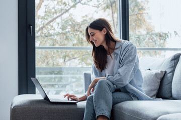 Concentrated millennial girl sit on couch working on laptop browsing internet at home during lazy weekend, focused young woman freelancer busy using computer surfing wireless web shopping online.