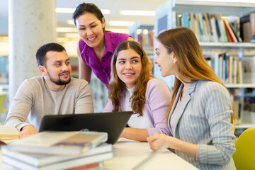 Obraz premium Group of enthusiastic young adult female and male colleagues working together in public library, discussing while sitting at table with laptop and books..