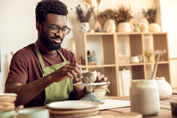 Dark-skinned man in eyeglasses shaping mug from clay and looking focused
