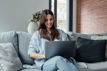 Concentrated millennial girl sit on couch working on laptop browsing internet at home during lazy...