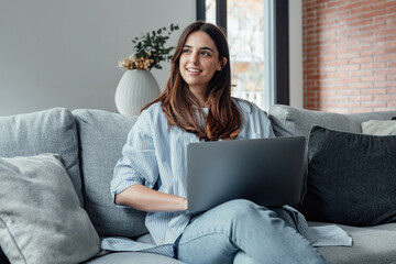 Concentrated millennial girl sit on couch working on laptop browsing internet at home during lazy...