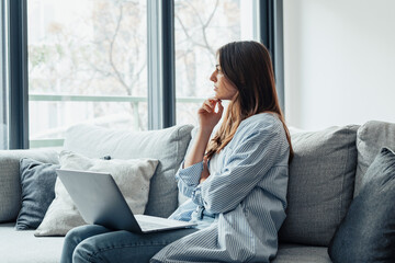 Distracted from work worried young woman sitting on couch with laptop, thinking of problems. Pensive unmotivated lady looking at window, feeling lack of energy, doing remote freelance tasks at home..