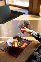 A person sits at a table in a restaurant, holding a glass of red wine and a plate of food