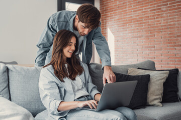 Happy millennial couple sit relax on couch in living room, smiling young husband standing from back and wife rest on sofa at home browsing Internet using modern computer device.