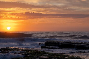 Fototapeta premium Bright orange sunrise beach waves crashing green mossy rocks clouds