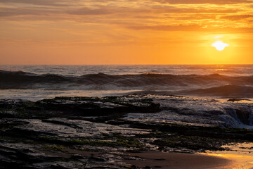Obraz premium Bright orange sunrise beach waves crashing green mossy rocks clouds