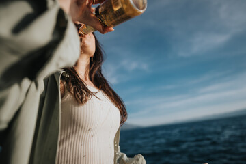 A joyful girl is drinking from a bottle, feeling happy and refreshed by the ocean under the clear blue sky.