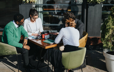 Diverse group of business colleagues in casual meeting at an outdoor cafe, focused on strategizing for company success.
