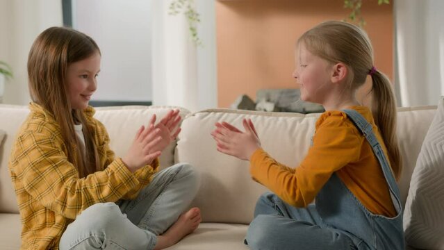 Happy playful Caucasian children little girls daughters siblings playing patty cake enjoy funny game together. Kids sisters fooling around clapping hands smile family sofa couch room relaxing holidays