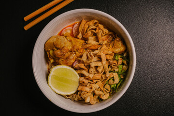 A bowl of Asian noodle dish with slices of chicken, lime wedge, and crispy noodles. The famous Thai noodle soup.
