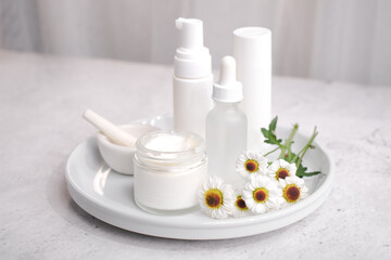 A collection of skincare products on a white tray, including a jar of cream, two pump bottles, and a smaller bottle, all in white packaging. Three daisy-like flowers add a touch of natural beauty.