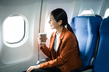 A woman in a formal suit sits in a window seat of an economy class section, looking out the...