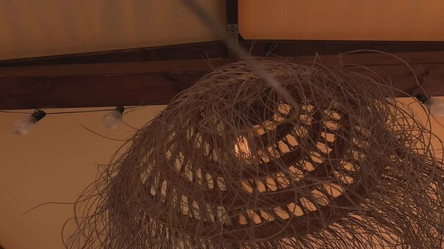 Incandescent light bulb lit inside the shell of the handmade lamp with reeds installed on the roof of the awning of the relaxation area at sea, shot traveling from below. Pontevedra, Galicia, Spain