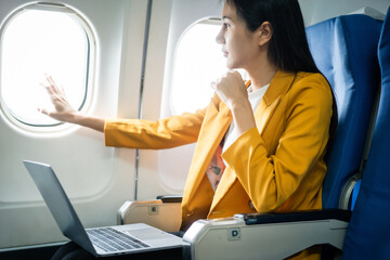 A young Asian woman, an airplane passenger, sits by the window seat, studying abroad. She gazes out the window, ready for takeoff in the economy class section.