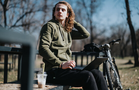 A young curly-haired man sits thoughtfully in a park, working on his laptop beside his bicycle, enjoying a sunny day outdoors.