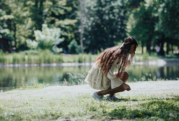 A beautiful young woman dressed in a light dress squats by a park lake, exuding a sense of freedom and joy on a sunny spring day.