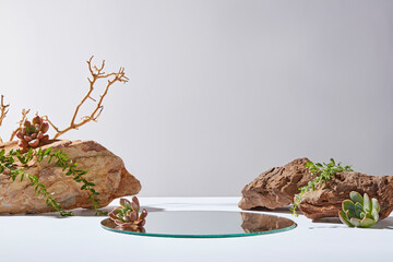 Light gray background photo from frontal shot, the display pedestal of glass placed on white countertop between two large brown rocks, dotted by green plants. Empty space for adding text
