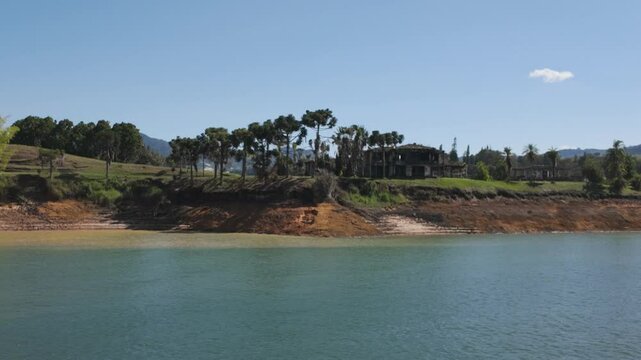 Ruined mansion of Pablo Escobar 'La Manuela,' located by lake in Colombia, with trees and clear blue sky - view from passing boat
