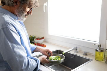 Mature man washing lettuce and vegetables in kitchen sink to prepare fresh salad for healthy lunch. Copy space.