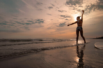 Surfer girl stretches and warms up her body before going surfing in the sea, on the sunset sky in the background