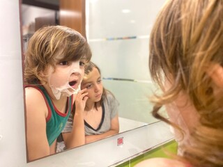 Young boy with a facial mask looking thoughtfully at his reflection in the bathroom mirror. He is...