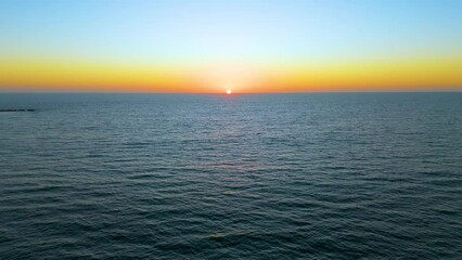 Experience the serene beauty of a tranquil aerial journey towards the sunset horizon over the sea, as the sun gently sets, casting warm, beautiful hues across the sky and water.