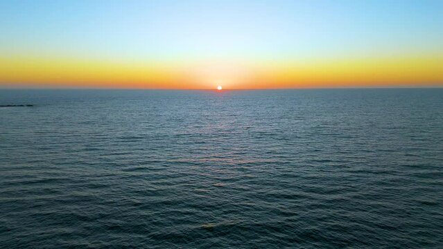 Experience the serene beauty of a tranquil aerial journey towards the sunset horizon over the sea, as the sun gently sets, casting warm, beautiful hues across the sky and water.