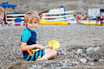 Adorable little blond kid boy building pebble stone castle on beach. Funny child playing with bucket and shovel. Vacations, summer, travel concept. Toddler enjoying summer vacations on sea.