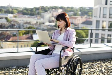 Business woman enjoys the view while working online on a roof top. Positive Caucasian lady in wheelchair sitting with laptop typing message while working remotely.