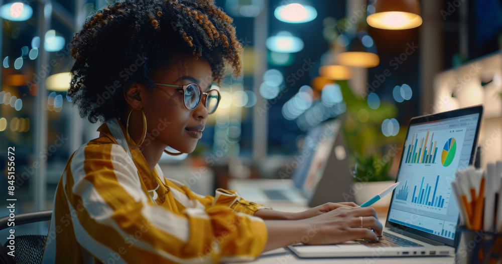 Poster a black female office worker working on a laptop at a desk in a modern office, looking at graphs and