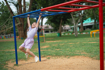 Asian young girl is hanging from a metal bar in a park. The scene is playful and fun, with the girl enjoying herself as she climbs the structure. The park is filled with other children and adults