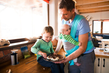 Father holding small baby in baby sling. Older brother eating porridge on kitchen counter....
