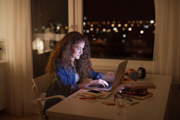 Beautiful university student is writing her thesis on a laptop at night, sitting at home at table, focusing on her research. Preparing for final exam, studying.