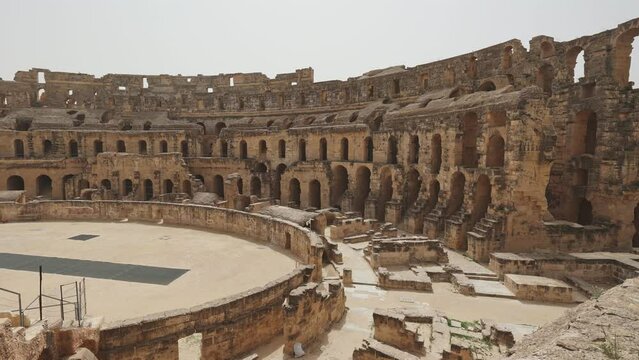 Scenic view of sun-drenched restored El Jem Amphitheatre arena encircled by ancient stone tiers and crumbling walls with archways under clear Tunisian sky. High quality 4k footage