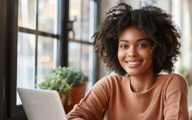 Fototapeta premium A woman with curly hair is smiling and sitting in front of a laptop. She is wearing a brown sweater and is looking directly at the camera