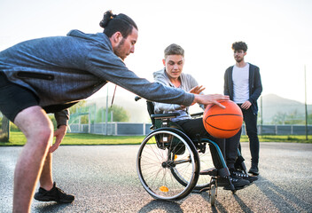 Obraz premium Disabled young man in a wheelchair playing basketball with his friends. Teamwork and male friendship.